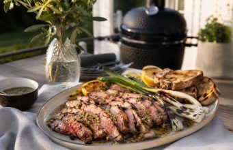 Pluma på grillen med sherryvinägrett och sotad salladslök Sliced medium-rare steak on a platter with lemon wedges, grilled bread, and asparagus, outdoors near a barbecue grill.