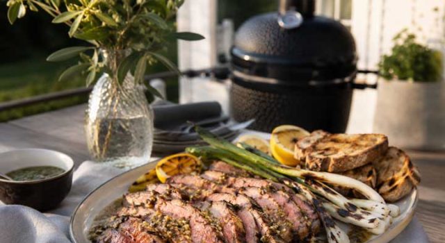Pluma på grillen med sherryvinägrett och sotad salladslök Sliced medium-rare steak on a platter with lemon wedges, grilled bread, and asparagus, outdoors near a barbecue grill.
