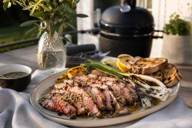Sliced medium-rare steak on a platter with lemon wedges, grilled bread, and asparagus, outdoors near a barbecue grill.