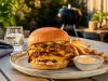 Double-stack cheeseburger with melted cheddar, onions, and pickles on a brioche bun, with fries and dipping sauce on an outdoor table.
