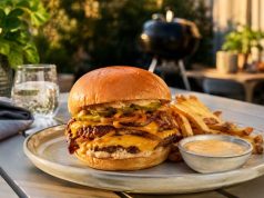 Double-stack cheeseburger with melted cheddar, onions, and pickles on a brioche bun, with fries and dipping sauce on an outdoor table.