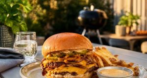 Double-stack cheeseburger with melted cheddar, onions, and pickles on a brioche bun, with fries and dipping sauce on an outdoor table.