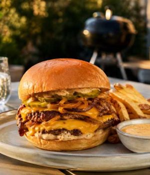 Double-stack cheeseburger with melted cheddar, onions, and pickles on a brioche bun, with fries and dipping sauce on an outdoor table.