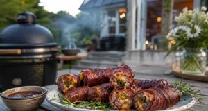 Plate of bacon-wrapped meat rolls with rosemary garnish and dipping sauce on a wooden table, outdoor grill in the background.