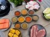 Assorted barbecue ingredients on a wooden table: salmon fillets, corn on the cob, raw steaks, chicken drumsticks, cabbage, and bowls of colorful spices with a grill nearby.
