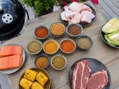 Assorted barbecue ingredients on a wooden table: salmon fillets, corn on the cob, raw steaks, chicken drumsticks, cabbage, and bowls of colorful spices with a grill nearby.