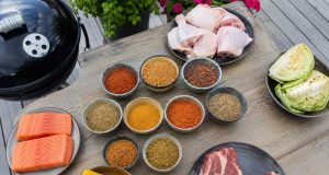 Assorted barbecue ingredients on a wooden table: salmon fillets, corn on the cob, raw steaks, chicken drumsticks, cabbage, and bowls of colorful spices with a grill nearby.