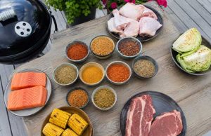 Assorted barbecue ingredients on a wooden table: salmon fillets, corn on the cob, raw steaks, chicken drumsticks, cabbage, and bowls of colorful spices with a grill nearby.