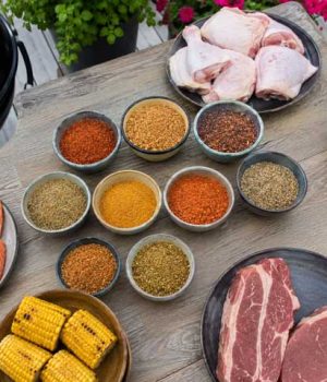 Assorted barbecue ingredients on a wooden table: salmon fillets, corn on the cob, raw steaks, chicken drumsticks, cabbage, and bowls of colorful spices with a grill nearby.
