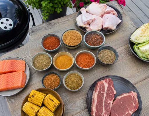Topp 10 BBQrubs du enkelt gör själv hemma Assorted barbecue ingredients on a wooden table: salmon fillets, corn on the cob, raw steaks, chicken drumsticks, cabbage, and bowls of colorful spices with a grill nearby.
