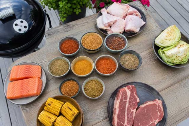 Assorted barbecue ingredients on a wooden table: salmon fillets, corn on the cob, raw steaks, chicken drumsticks, cabbage, and bowls of colorful spices with a grill nearby.