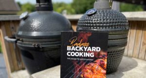 Paperback cookbook titled 'Badass Backyard Cooking' rests on a concrete railing with two smokers in the background.