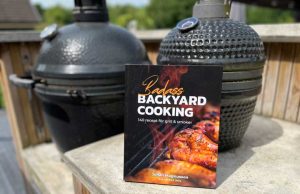 Paperback cookbook titled 'Badass Backyard Cooking' rests on a concrete railing with two smokers in the background.