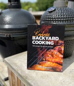 Badass-backyard-cooking Paperback cookbook titled 'Badass Backyard Cooking' rests on a concrete railing with two smokers in the background.