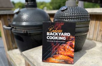 Paperback cookbook titled 'Badass Backyard Cooking' rests on a concrete railing with two smokers in the background.