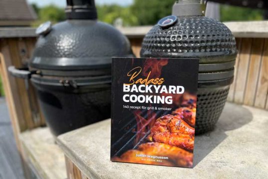 Paperback cookbook titled 'Badass Backyard Cooking' rests on a concrete railing with two smokers in the background.